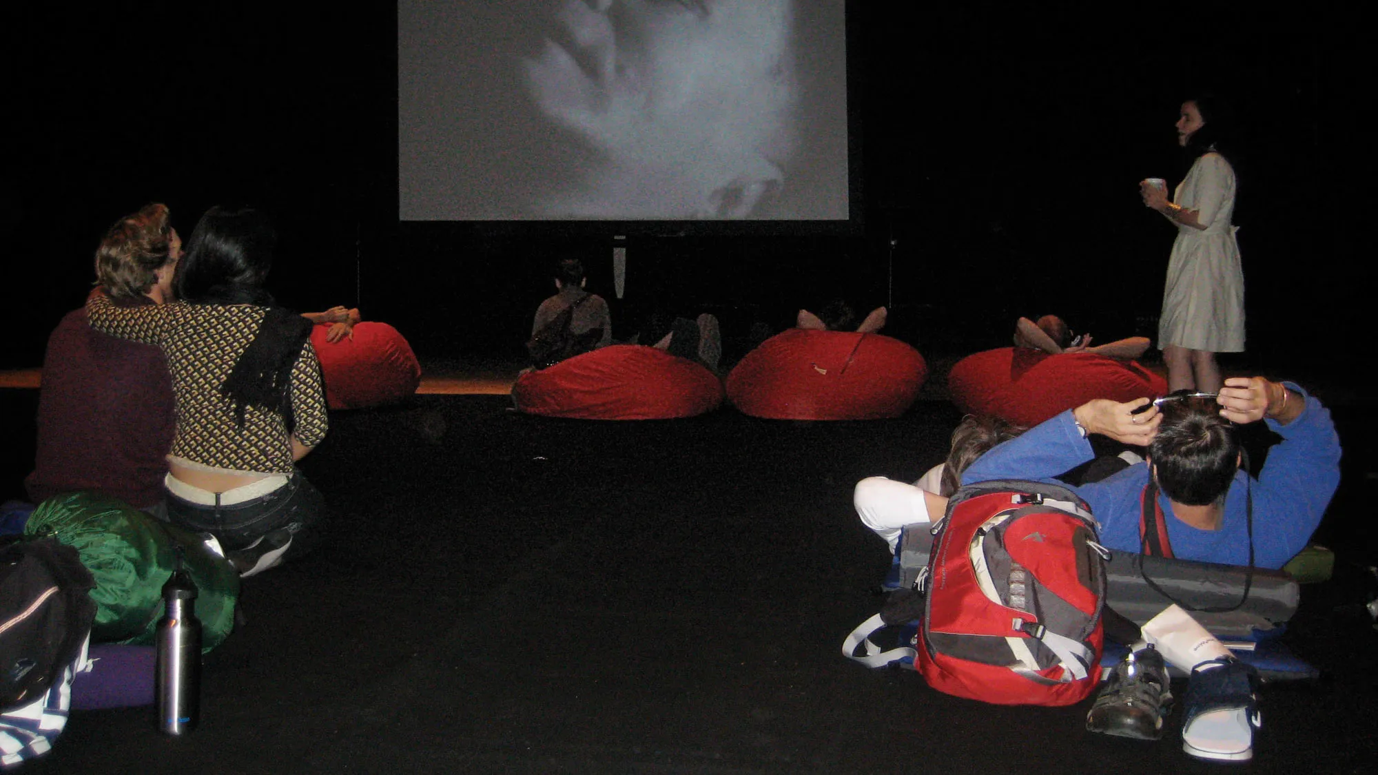 Various people seated in red bean bag chairs in a theater. A couple sits in the foreground in an embrace, a woman in a white dress finds her seat while holding a cup. 