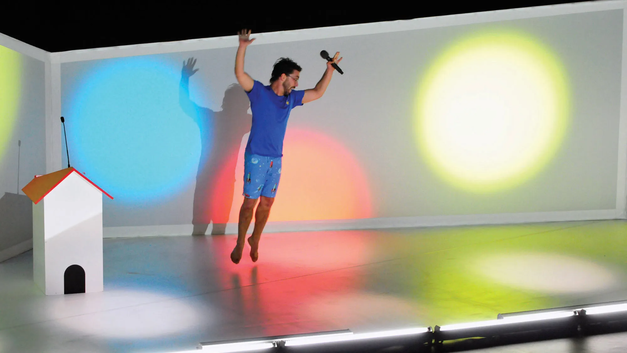 A man wearing a blue t-shirt and shorts jumping with arms up in a white room lit with primary colored lights.