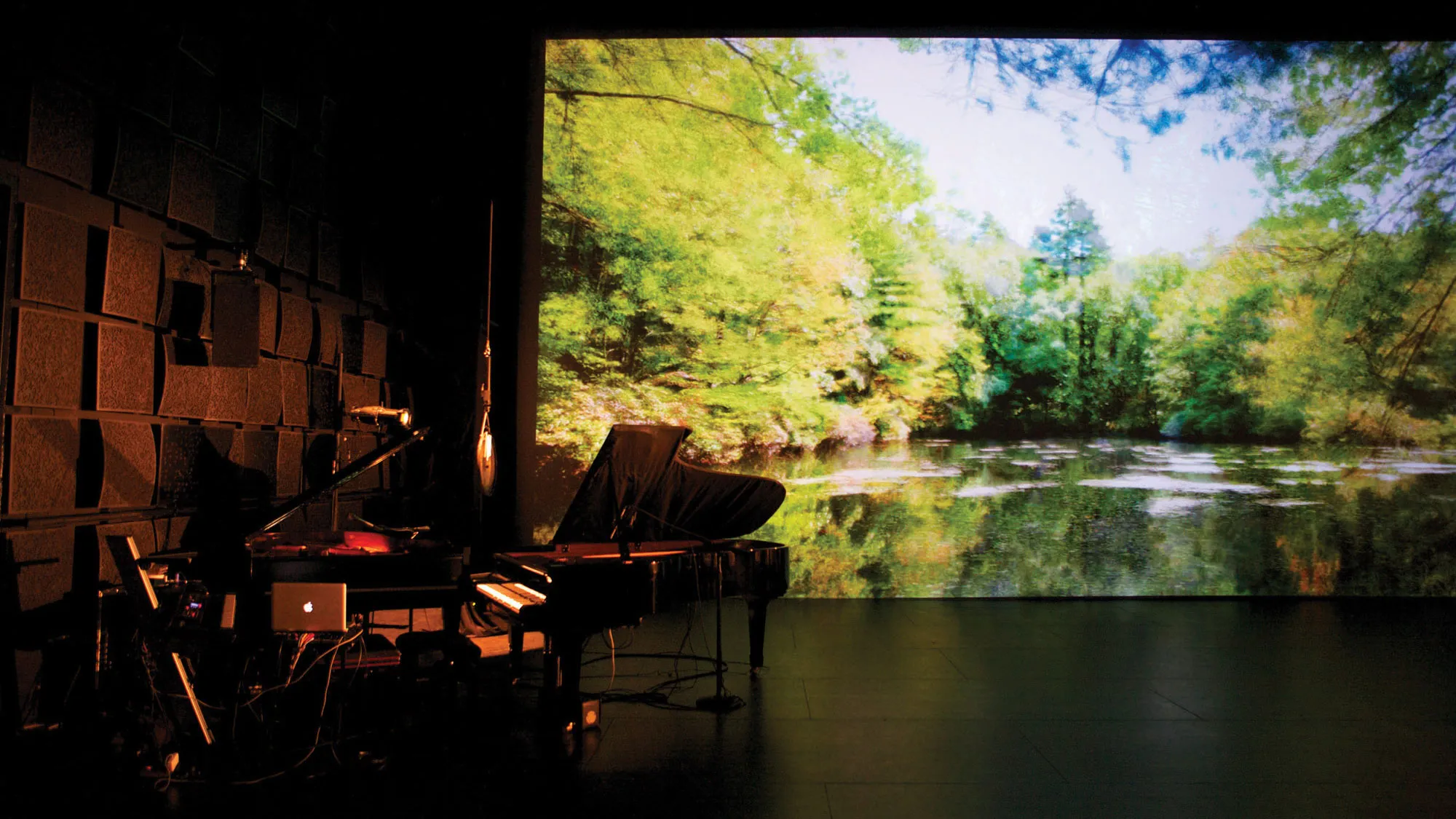 Two grand pianos among various wires, microphones, and recording devices in a black box studio in front of a projection of a tranquil green pond.
