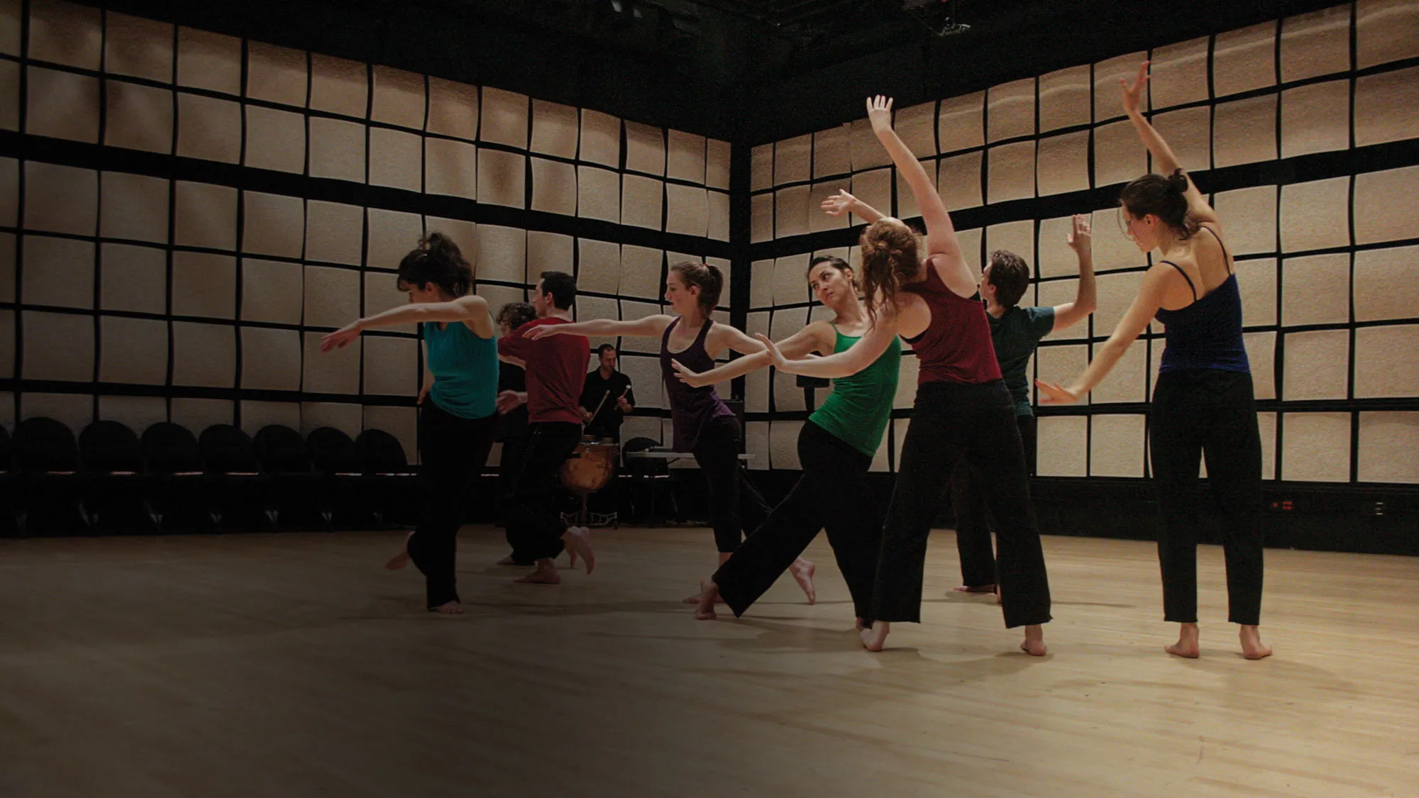 Eight dancers wearing jewel toned tops with black bottoms, moving with arms outstreched in a room with tan acoustic tiles on the walls. 