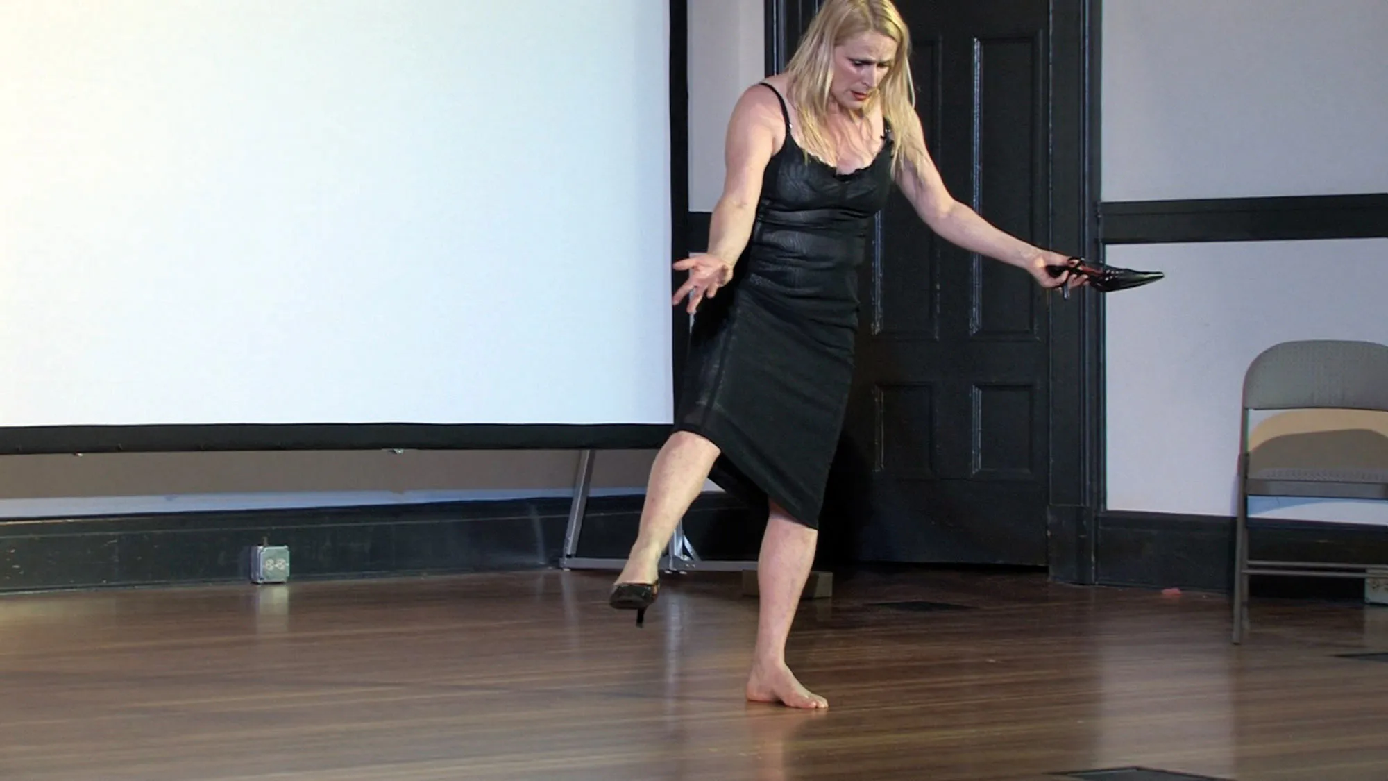 A femme dancer wearing a black slip dress and one black heel standing on the shoeless foot with the other leg extended and clutching the missing black heel. She looks down with hands out to the side.