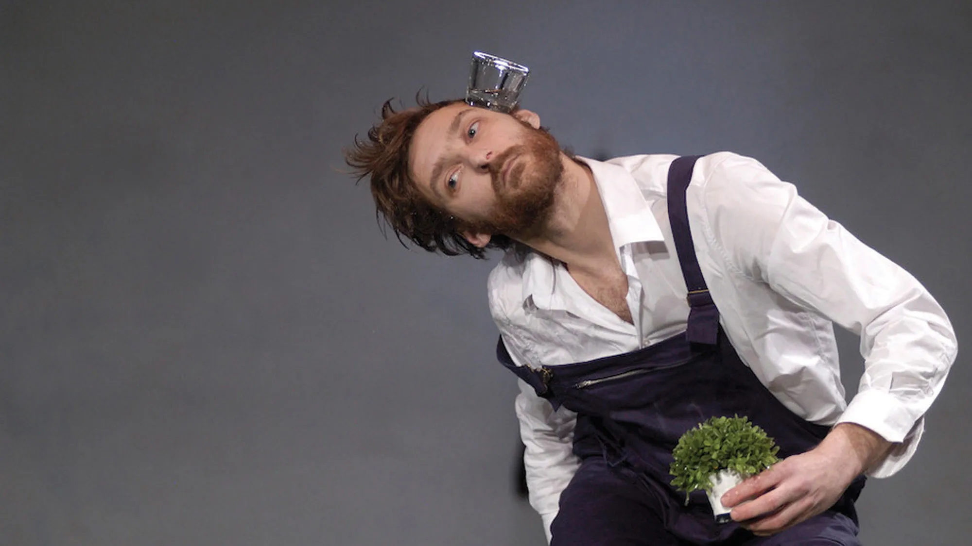 a man wearing dark gray overall and white shirt sitting holding a small plant while balancing a glass of water on the side of his head. 