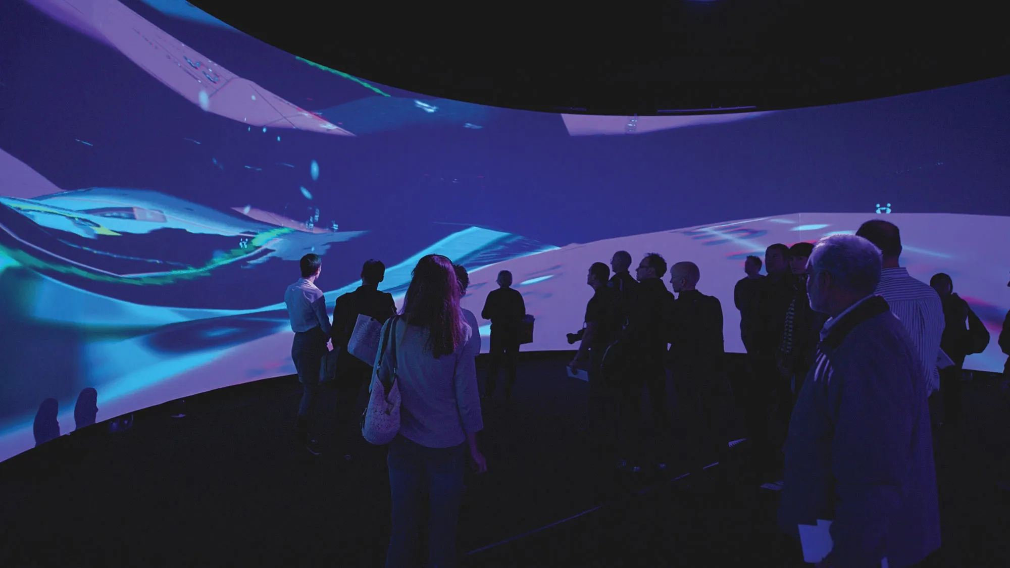 A small audience silhouetted against a panoramic screen showing abstract purple and teal waves. 