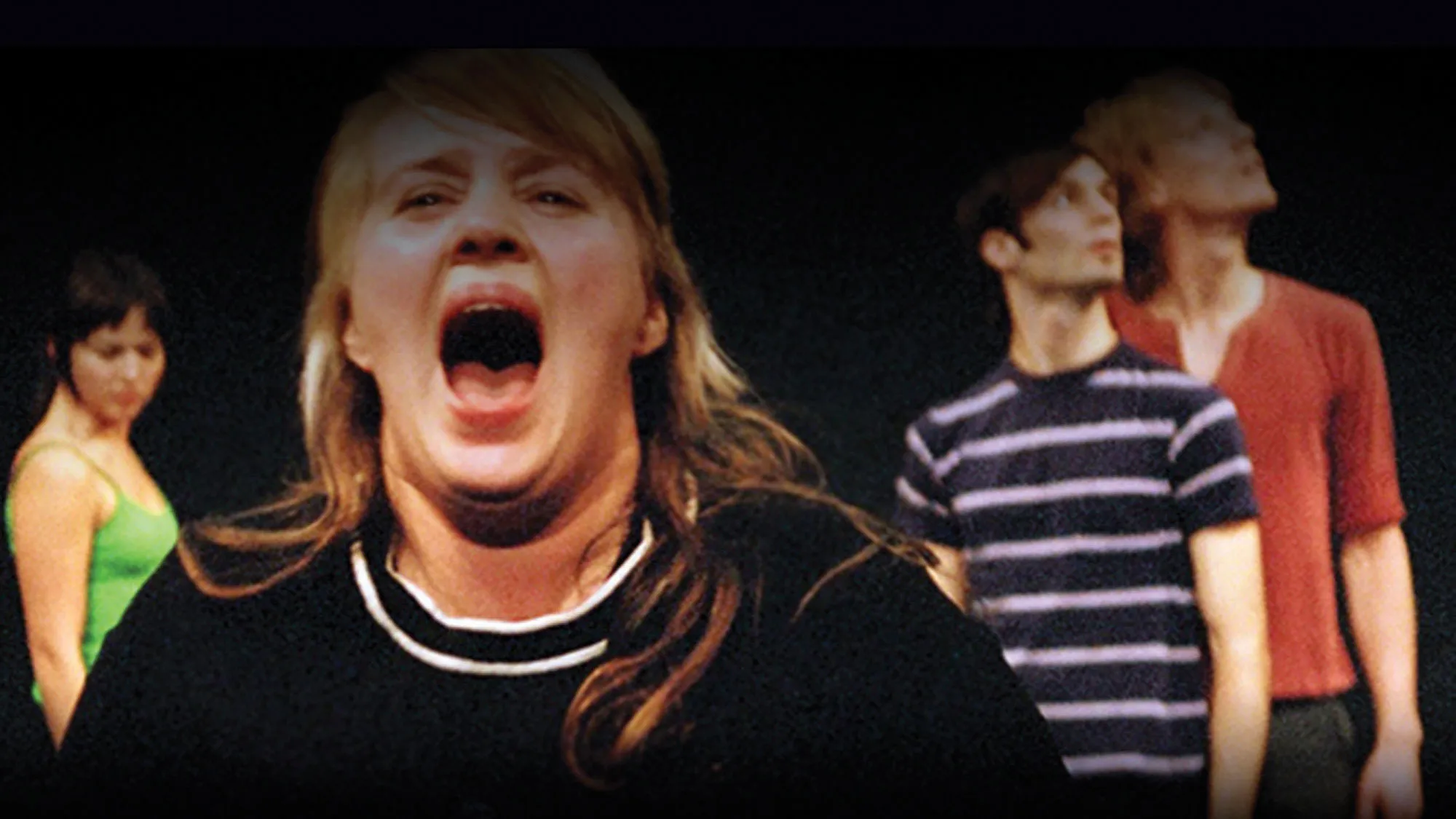 A white woman with blonde hear and wearing a black mock neckshirt screaming directly into the camera. Three people can be seen milling about int he background. 