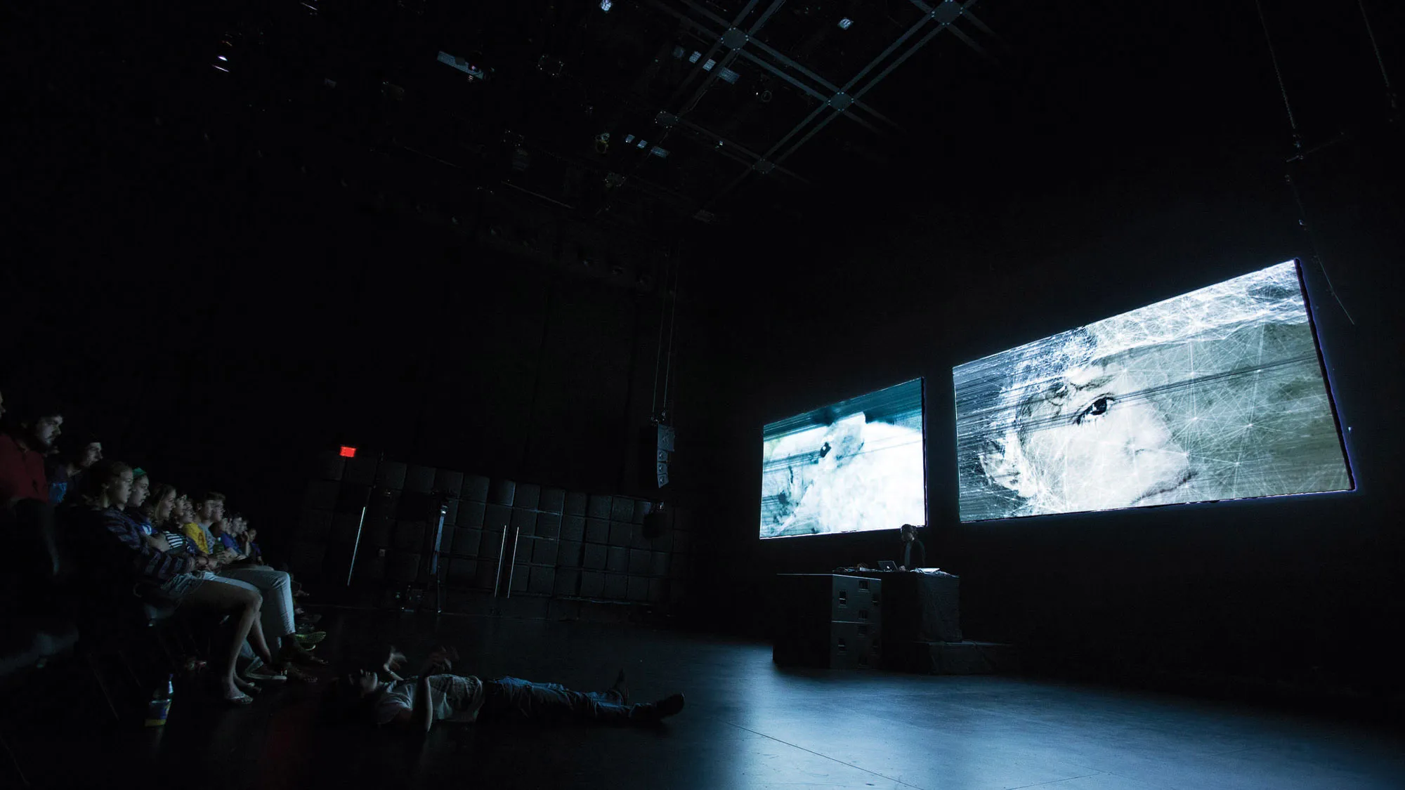 Two screens projecting scratched images of human eyes to a seated audience in a black theater. 