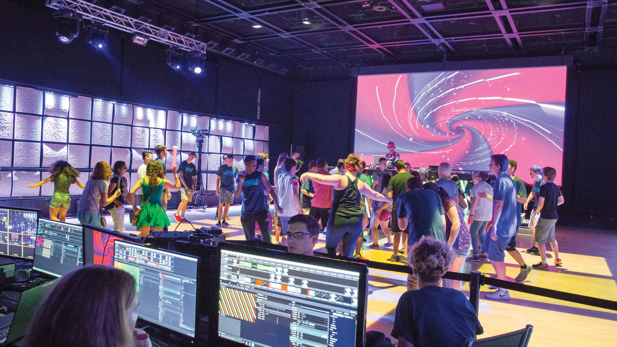 A crowd of people dancing on a yellow dance floor in front of a projection of a pink and purple swirl. 