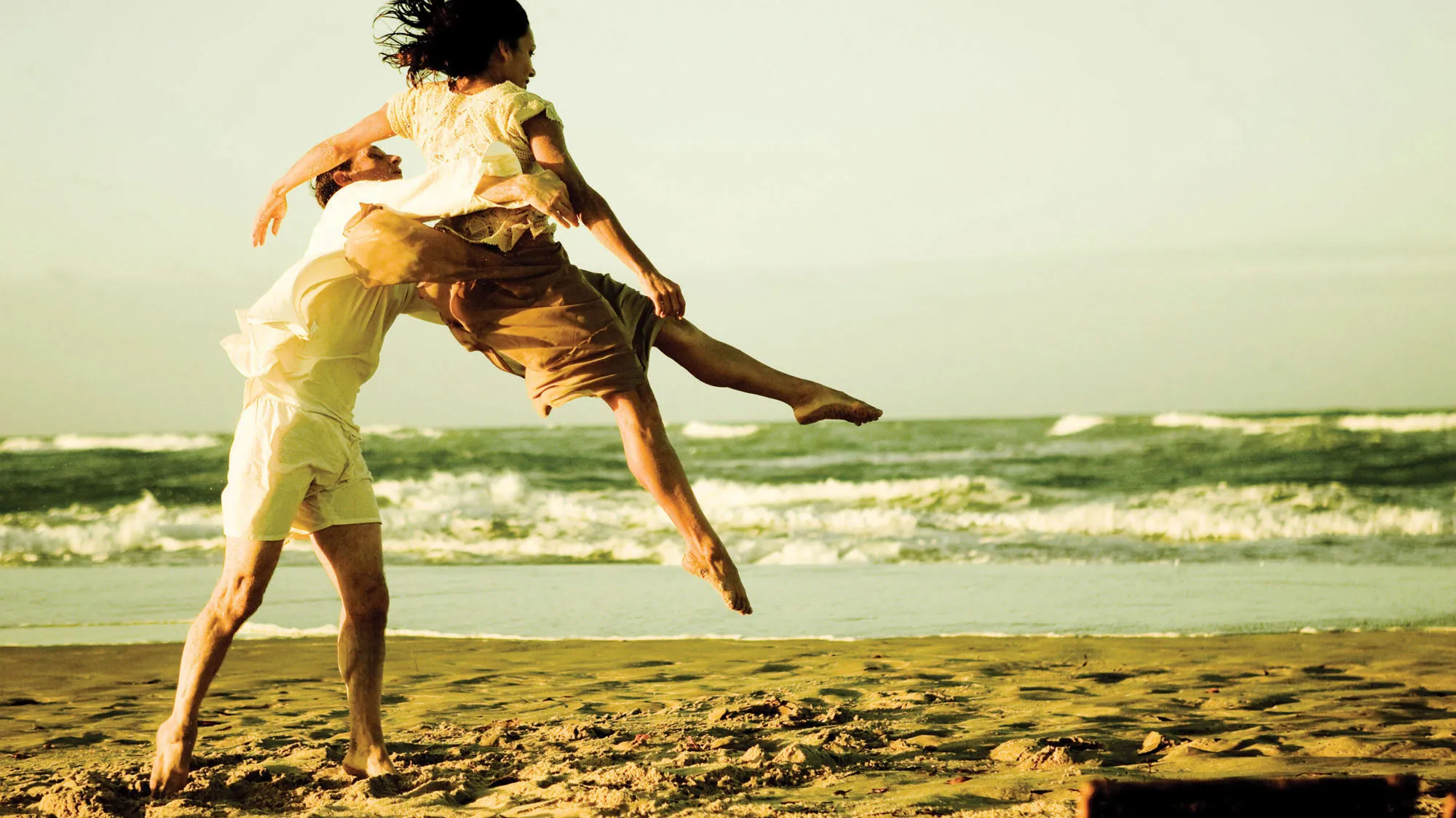 A man dressed casually tosses a woman into the air on a beach lit by warm yellow light. 