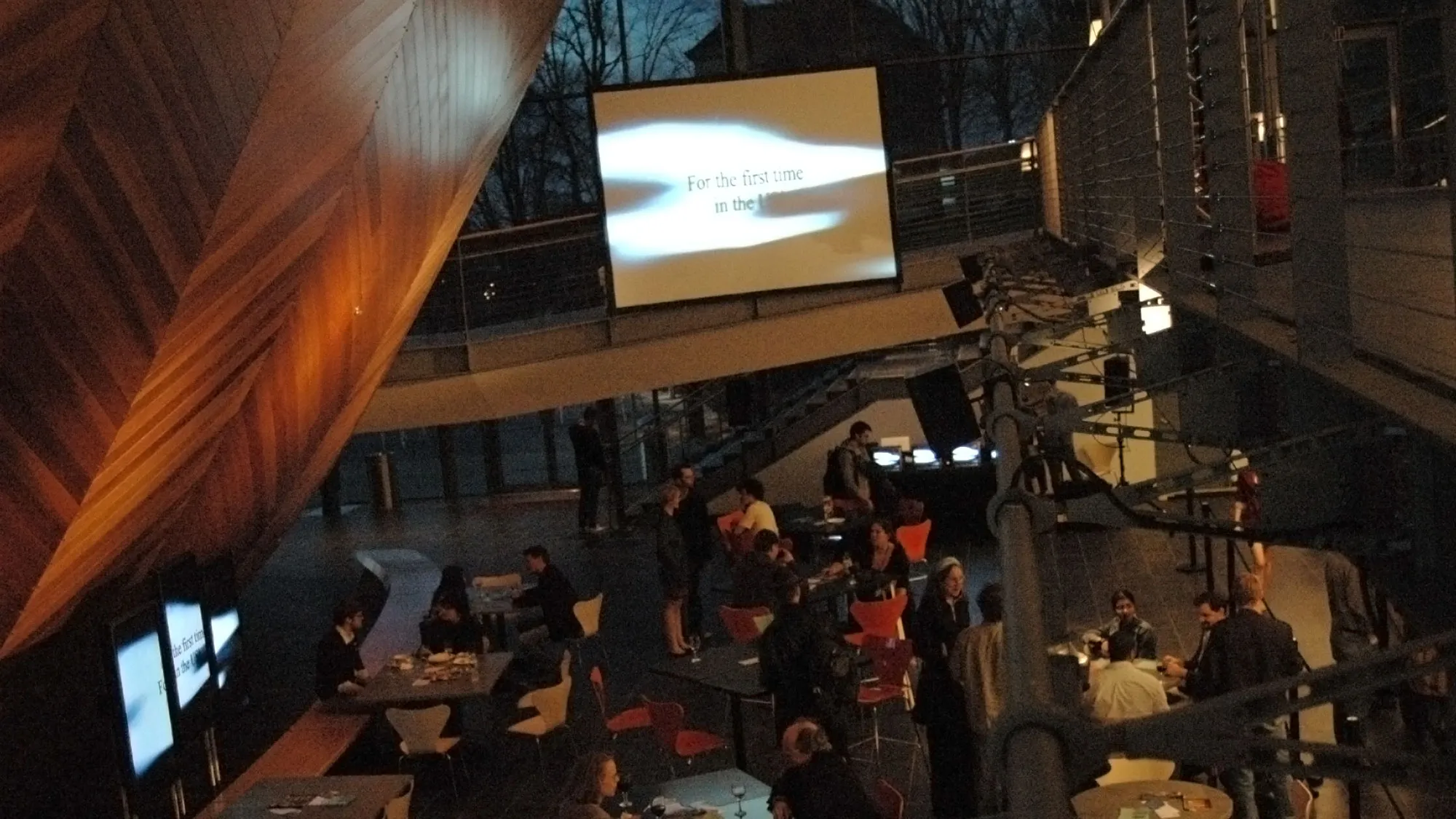 A reception full of people milling about under the hull of the EMPAC concert hall. A screen projecting the words "For the first time in the.." is suspended above the crowd.