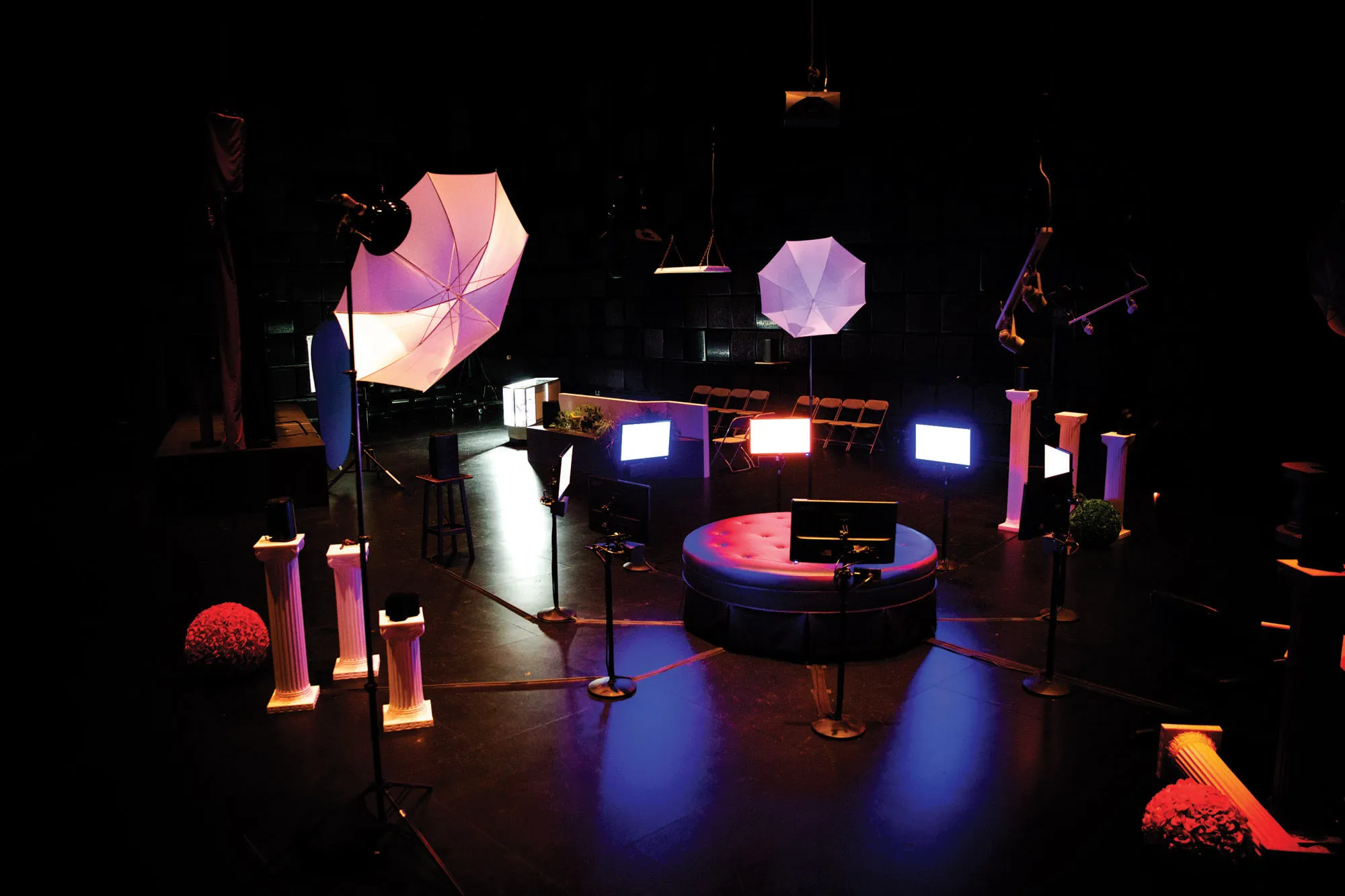 Behind the scenes, showing a round tufted bed surrounded by various lighting rigs and decorative columns washed in pink light. 