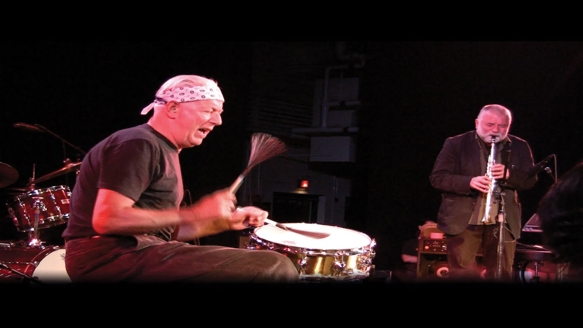 A white man wearing a bandana tied around his head playing the drums alongside another white man with a gray beard playing soprano sax. 