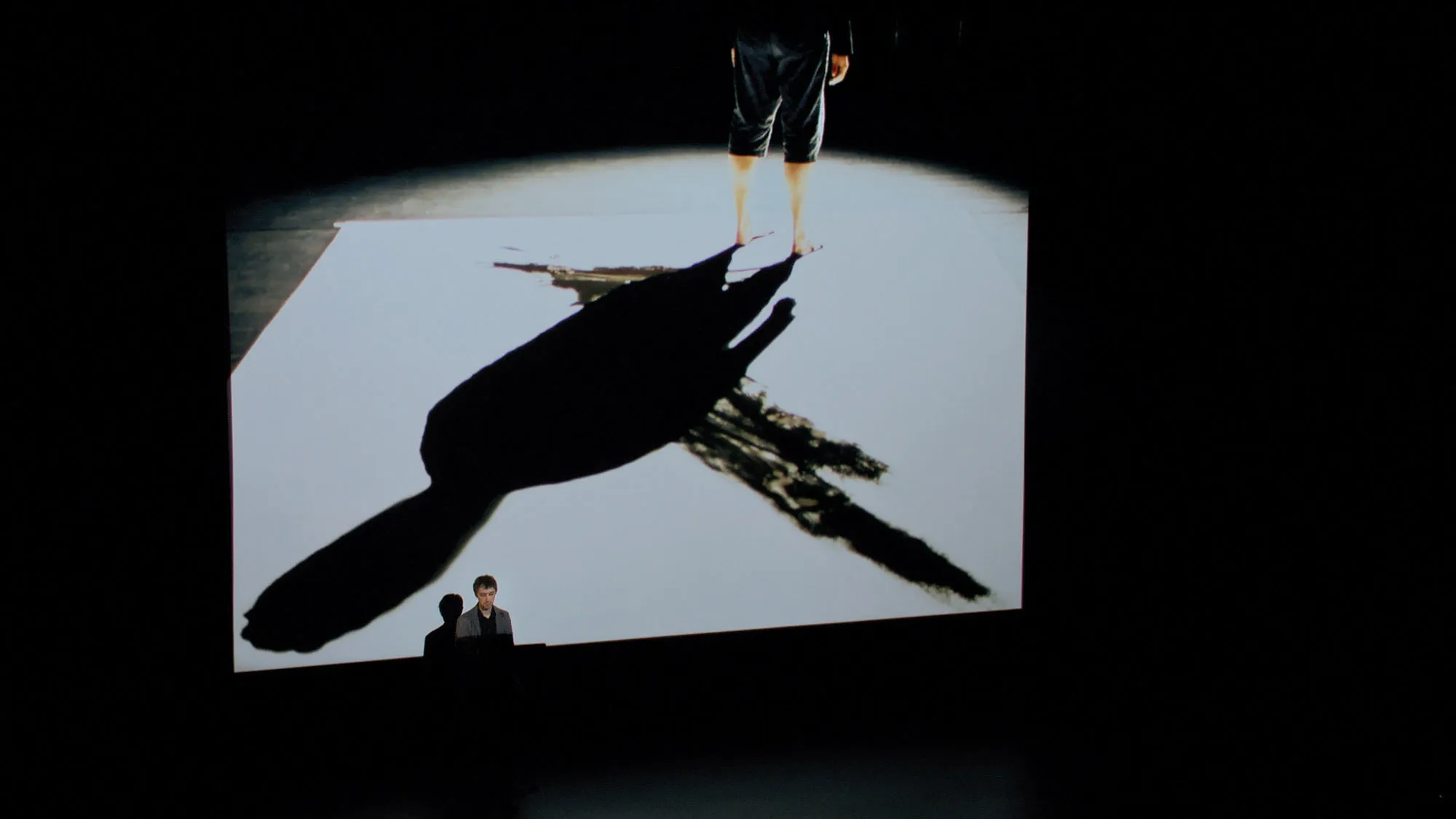 A large screen displaying the shadow of a man wearing shorts and standing in the upper right corner. 