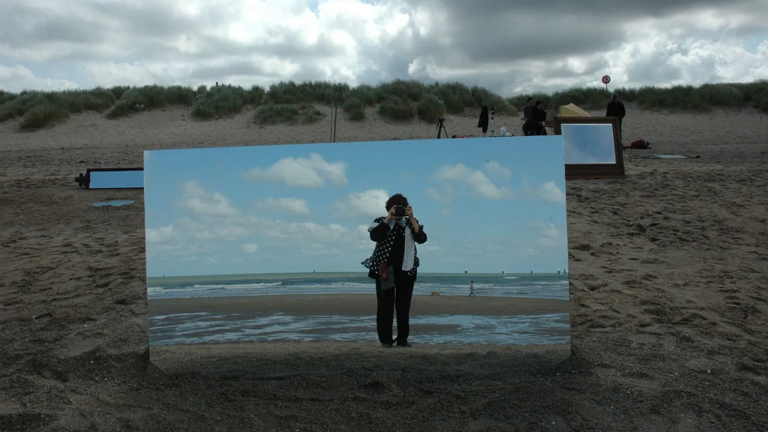 Agnes Varda taking a picture of herself in a large mirror placed on a beach.