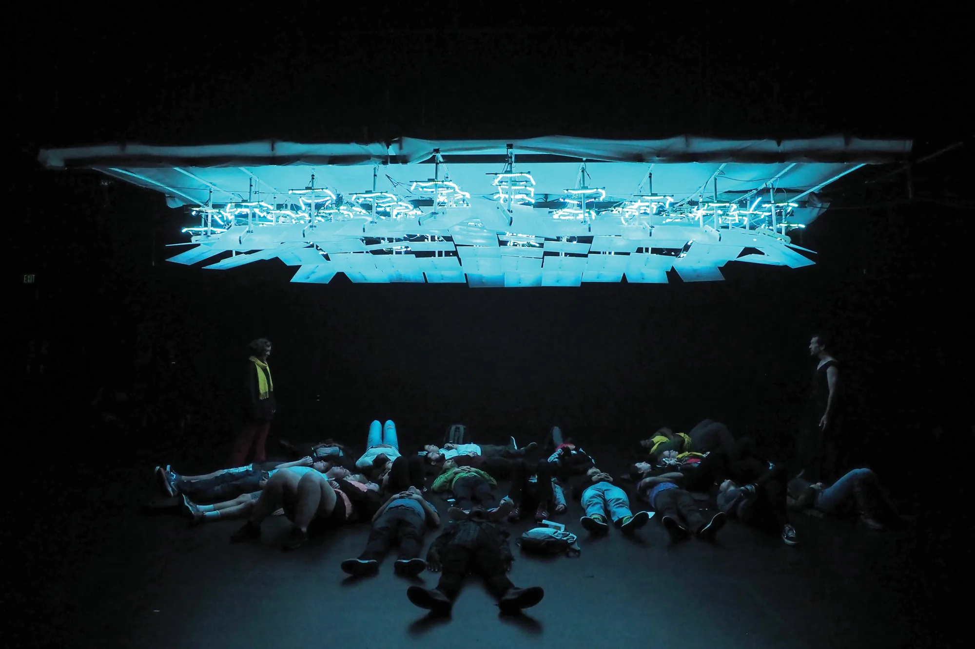 a group of about 15 people laying under a grid of lighted squares on the theatre stage