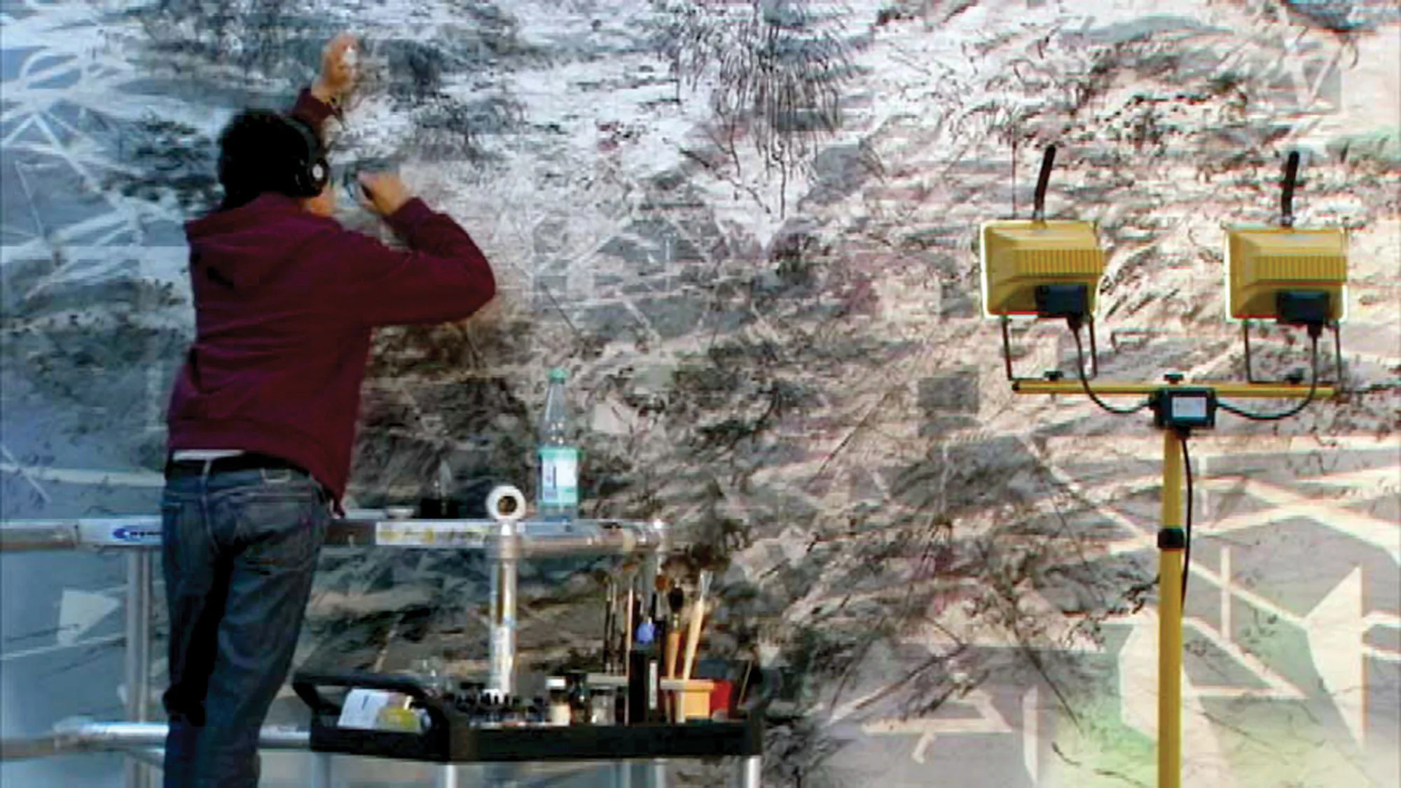 A man wearing a burgundy hoodie and jeans leans against a wall covered in an abstract expressionist style painting in gray, white, and black. 