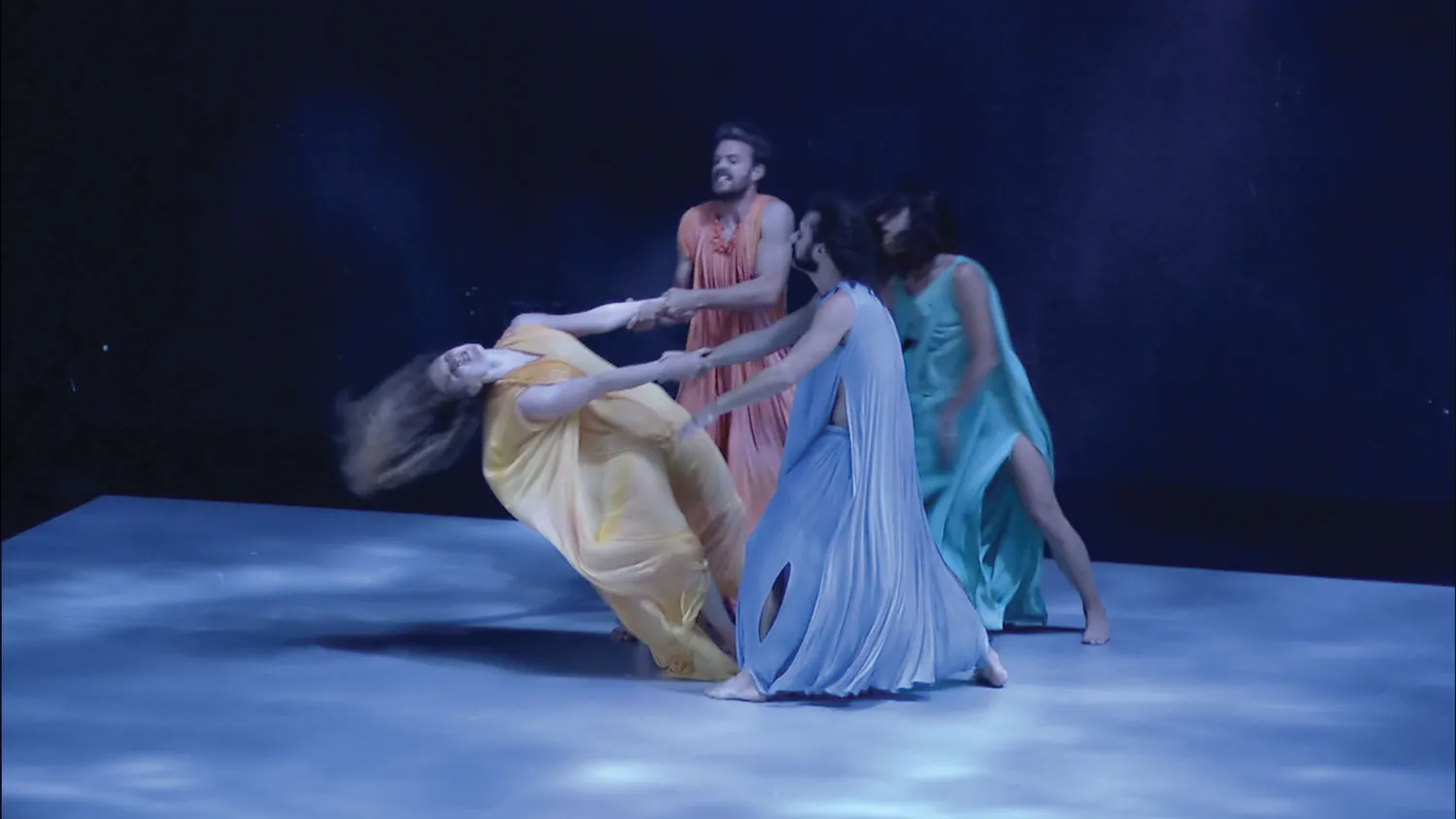 Four dancers wearing flowy pastel costumes dancing in a circle. A woman in yellow throws herself back dramatically as the other three grab her arms. 