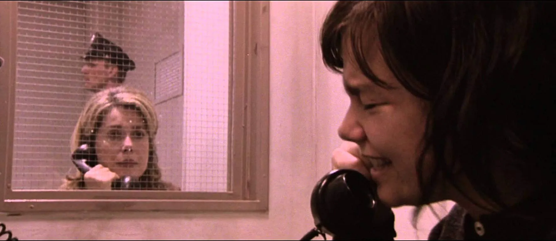 A  woman crying into the phone as another sitting behind prison glass listens. A guard stands in the background. 