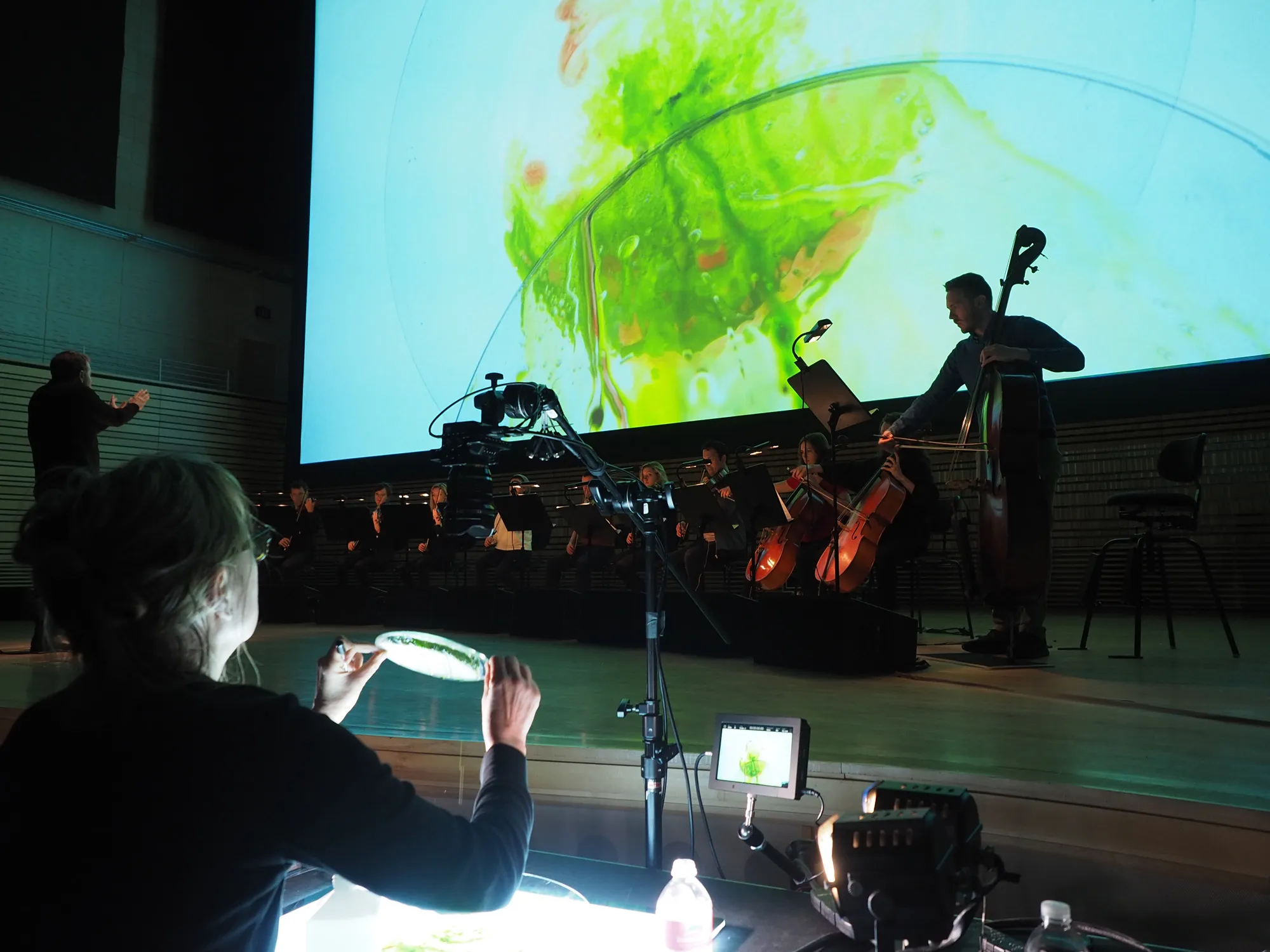 An orchestra on stage in front of a projected image of an lime green blob that is being controlled by a woman in the foreground.