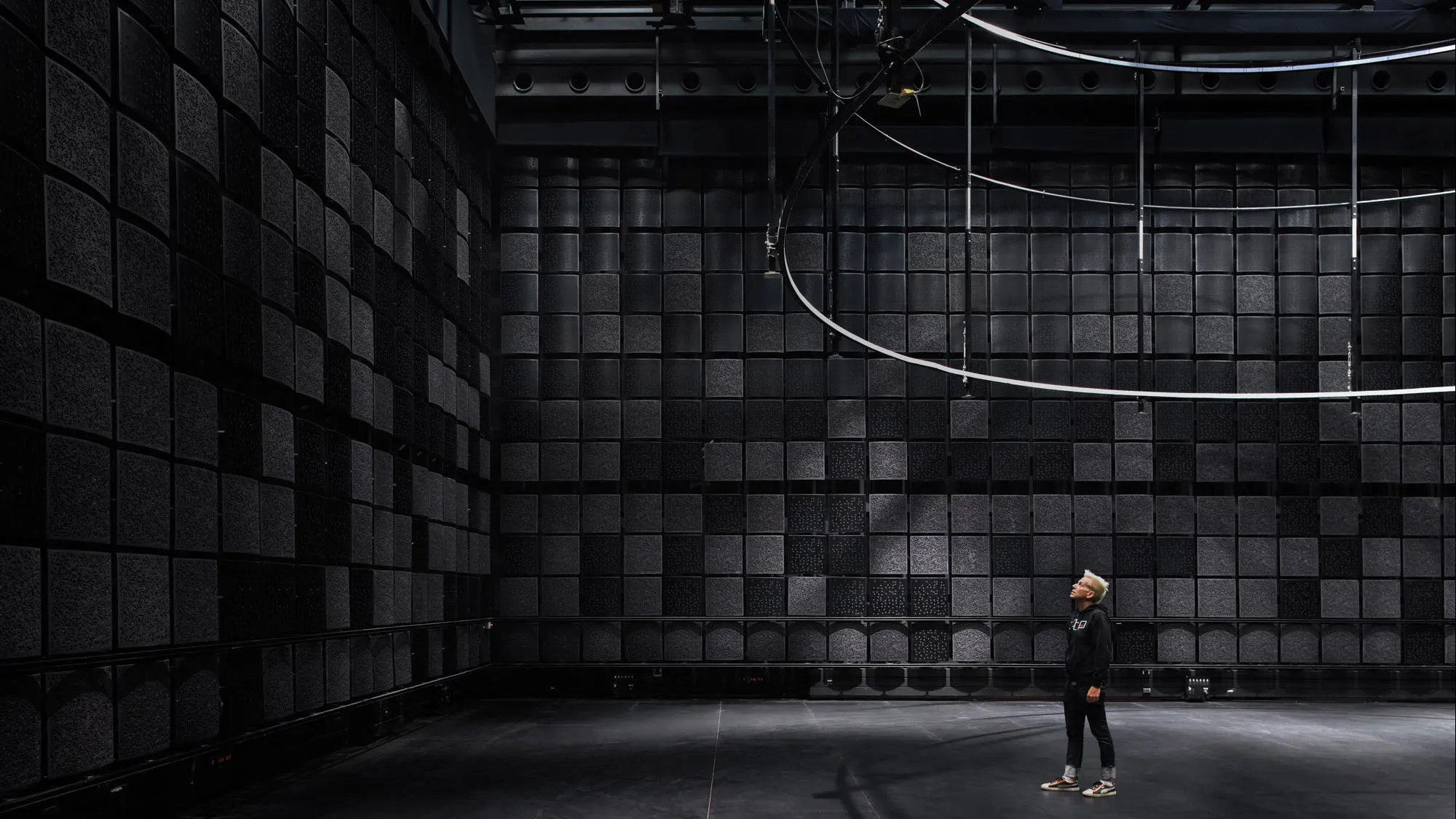 A man in a room full of black acoustic tiles with a rigging apparatus hanging above.  