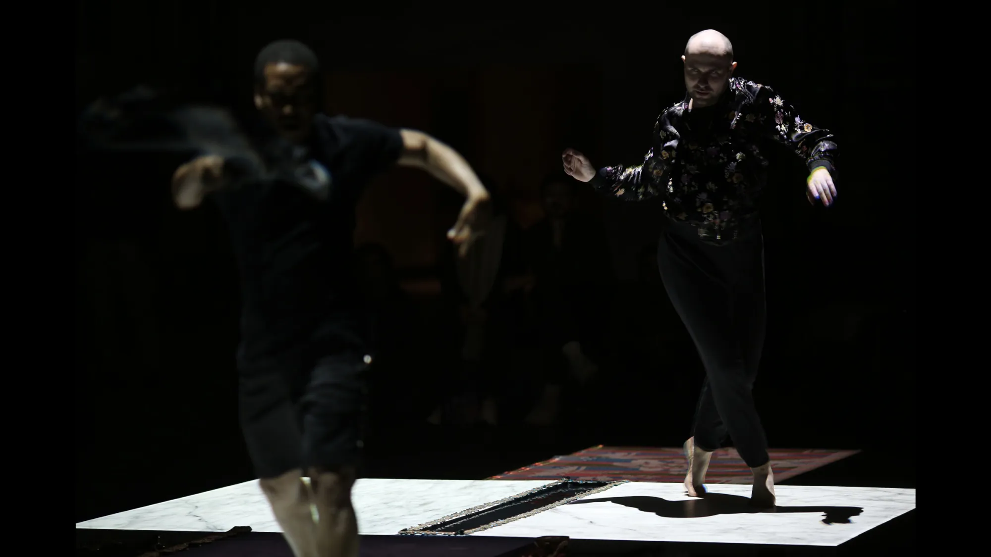 Two male dancers voguing across a stage light by squares of white light on the ground.
