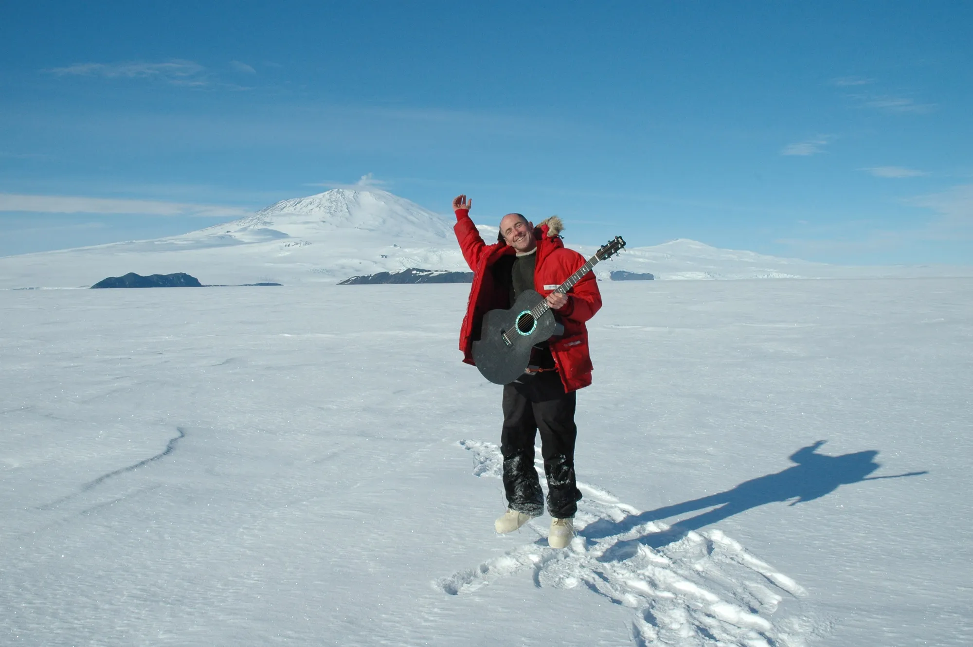 A white man wearing a red coat jumping in the snowy tundra of Antartica while play an acoustic guitar.