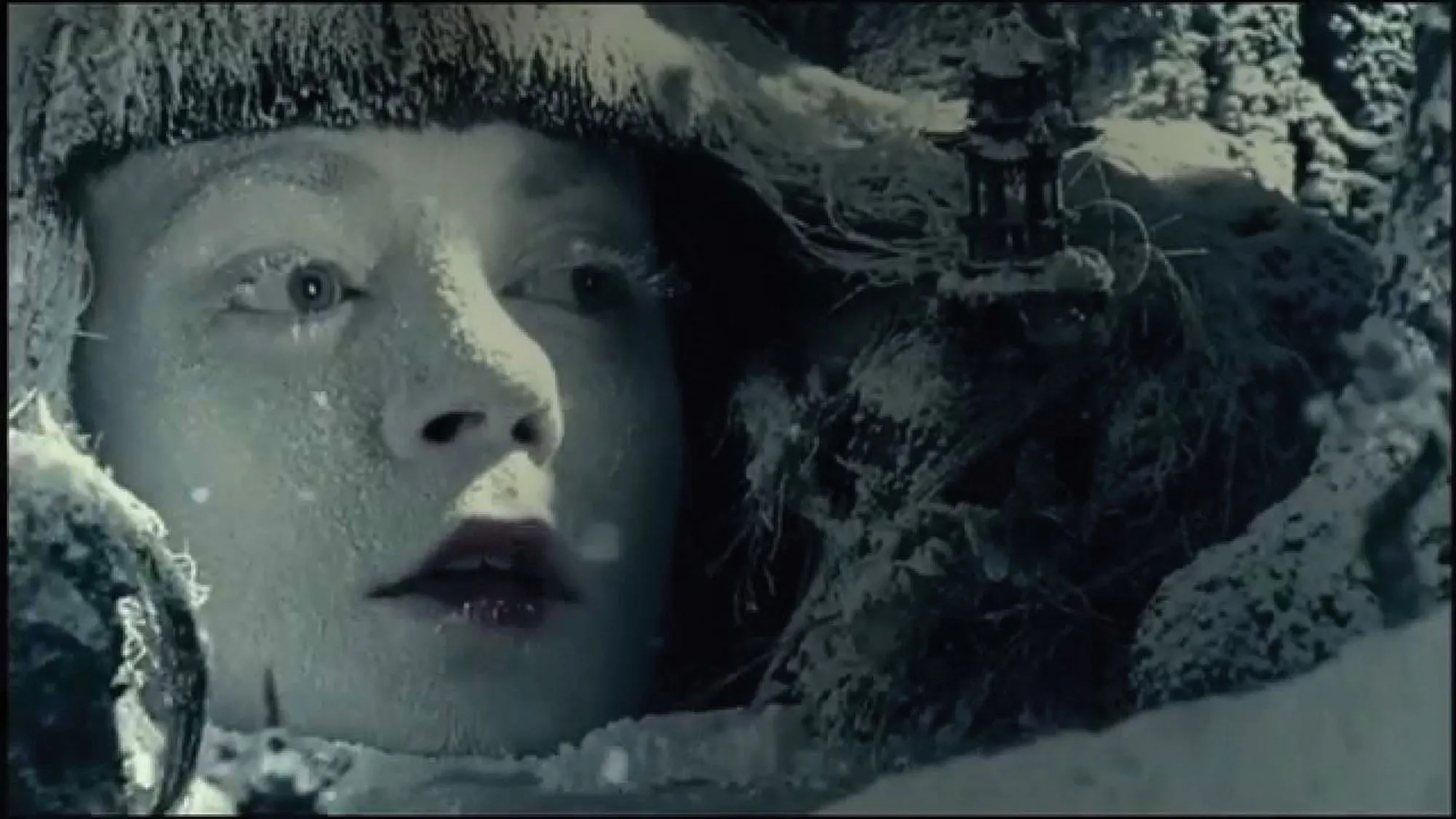 A woman's face painted white with red lips and frost on her eyelashes to evoke being frozen against a back drop of snowy pine trees.
