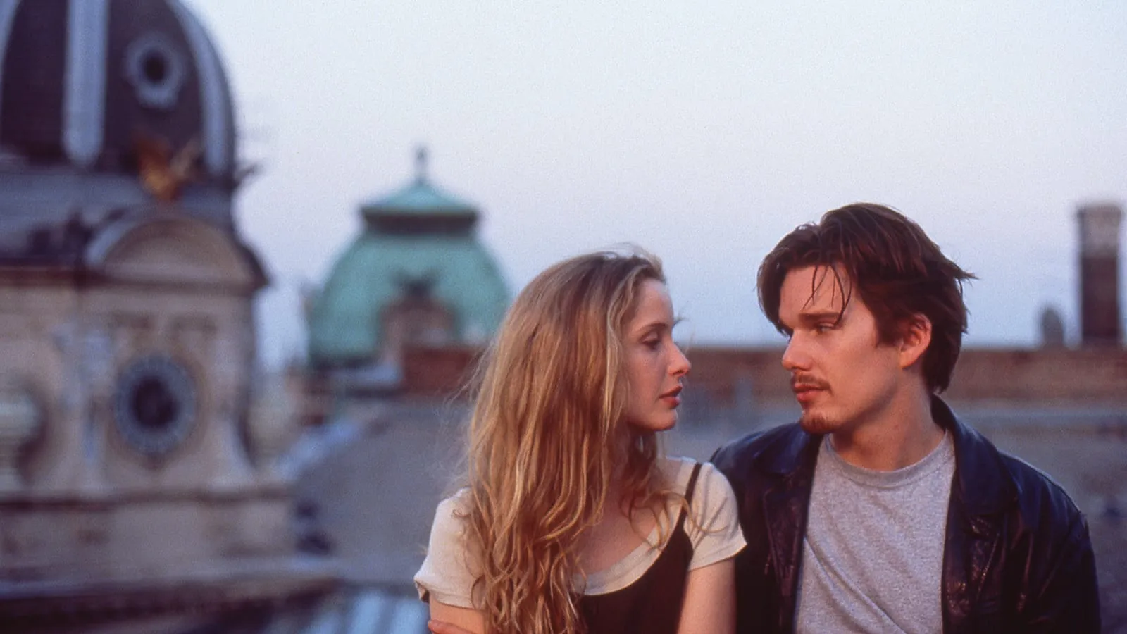 A woman with blonde hair and a man with brown hair looking longingly into each other eyes while sitting on a roof top overlooking a European cityscape. 