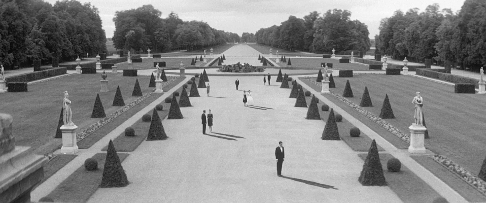 A structured English garden with a large boulevard lined by triangular shrubs, spherical bushes, and statues. Nine people pressed in formal attire mill about.