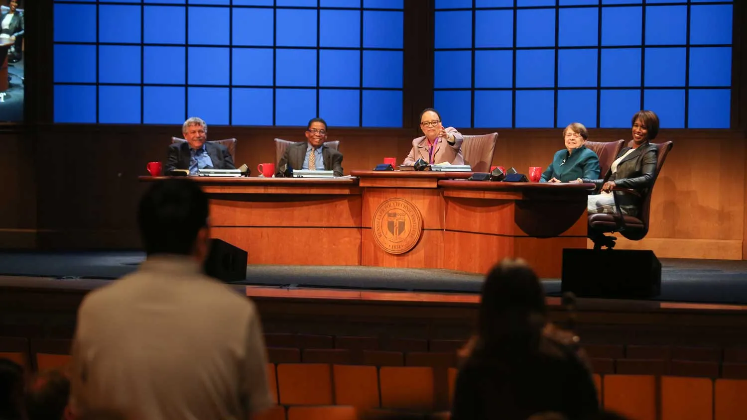 Dr. Shirley Ann Jackson and her colloquy guest panel seated behind a large wooden desk with the RPI seal. Behind them are projected images of a blue grid to evoke the idea of windows. Dr. Jackson points into the audience. 