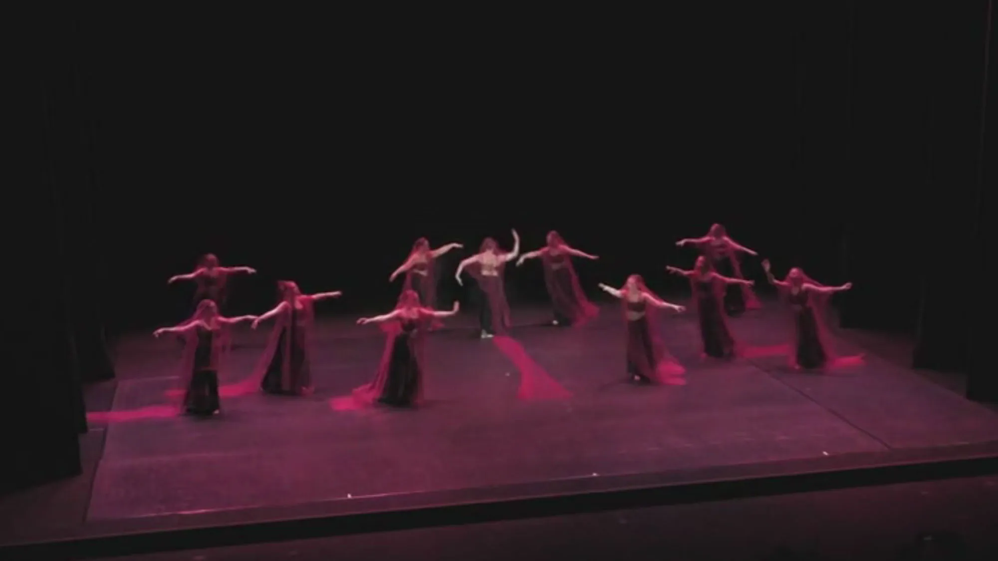 Eleven female dancers on stage wearing sheer red veils and dark dresses, moving towards the center of the pink lit stage with arms outstretched.  
