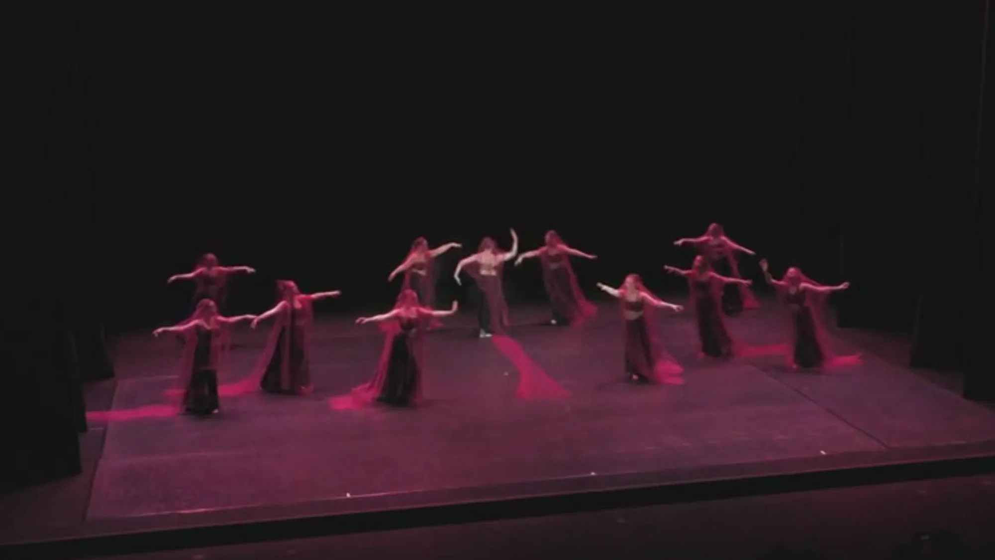 Eleven female dancers on stage wearing sheer red veils and dark dresses, moving towards the center of the pink lit stage with arms outstretched.