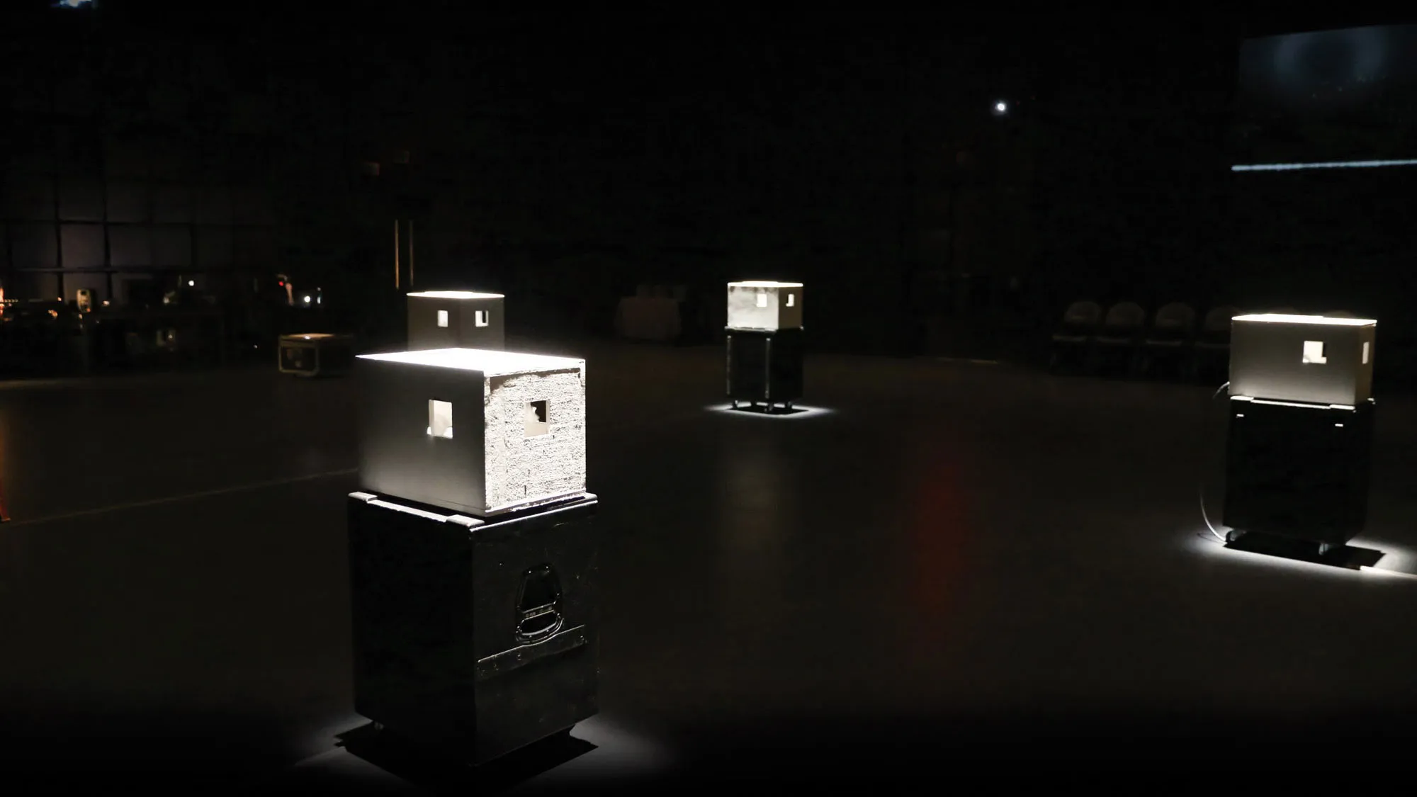 Four lit white boxes with small cutouts on black pedestals in a black box studio. 