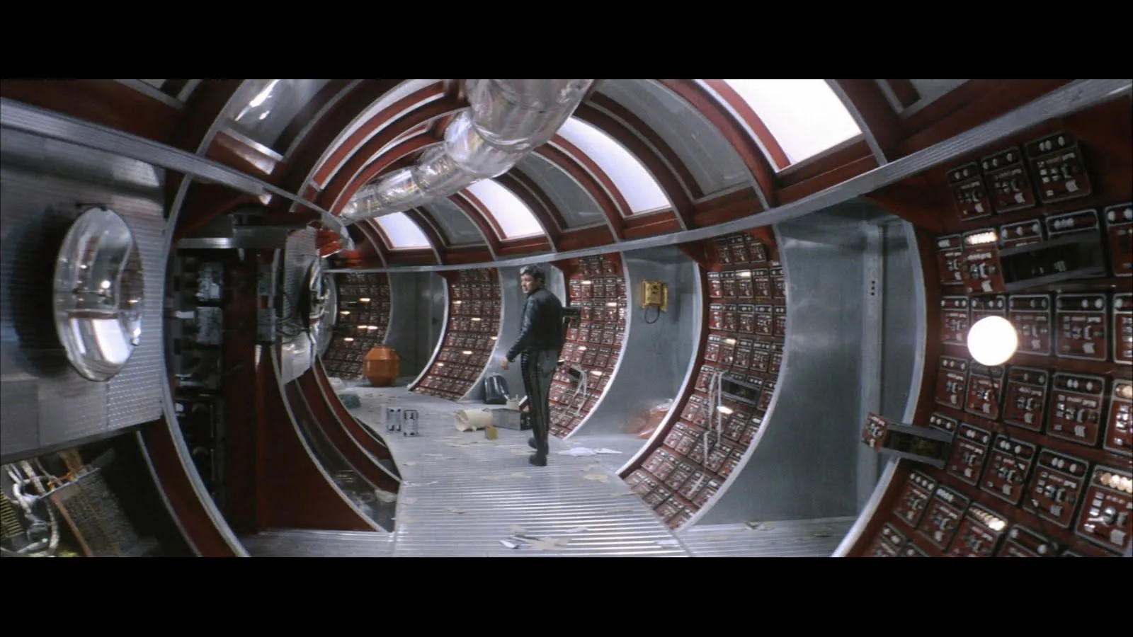 A man standing inside a messy hallway of a fictional spaceship. The steel gray and burgundy tunnel is lined in various nobs and buttons.