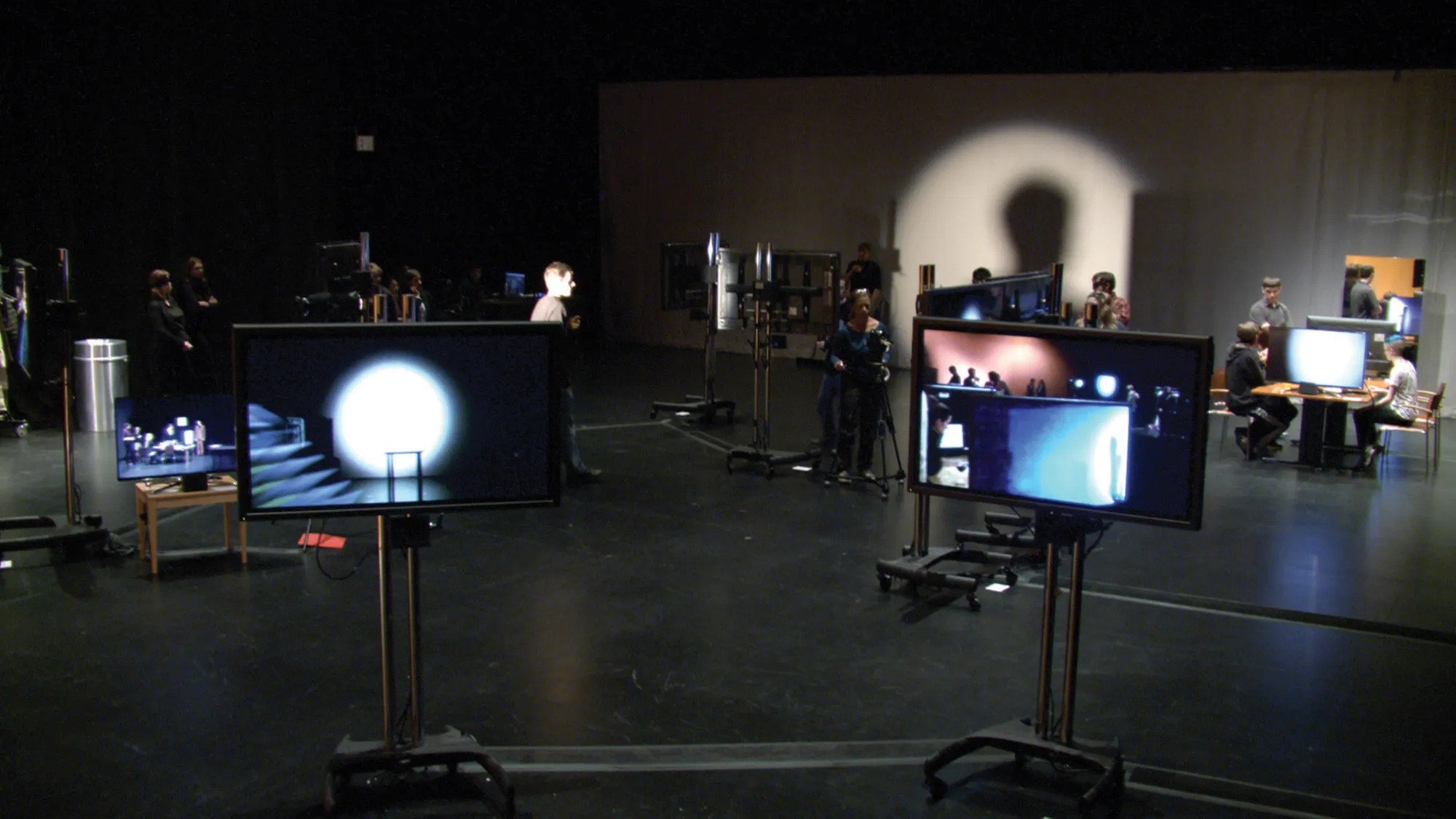 Two screens on legs in for the foreground showing images of the rest of the room. Various crew members work behind the scenes in front of a wall lit by a soft circle of white light. 