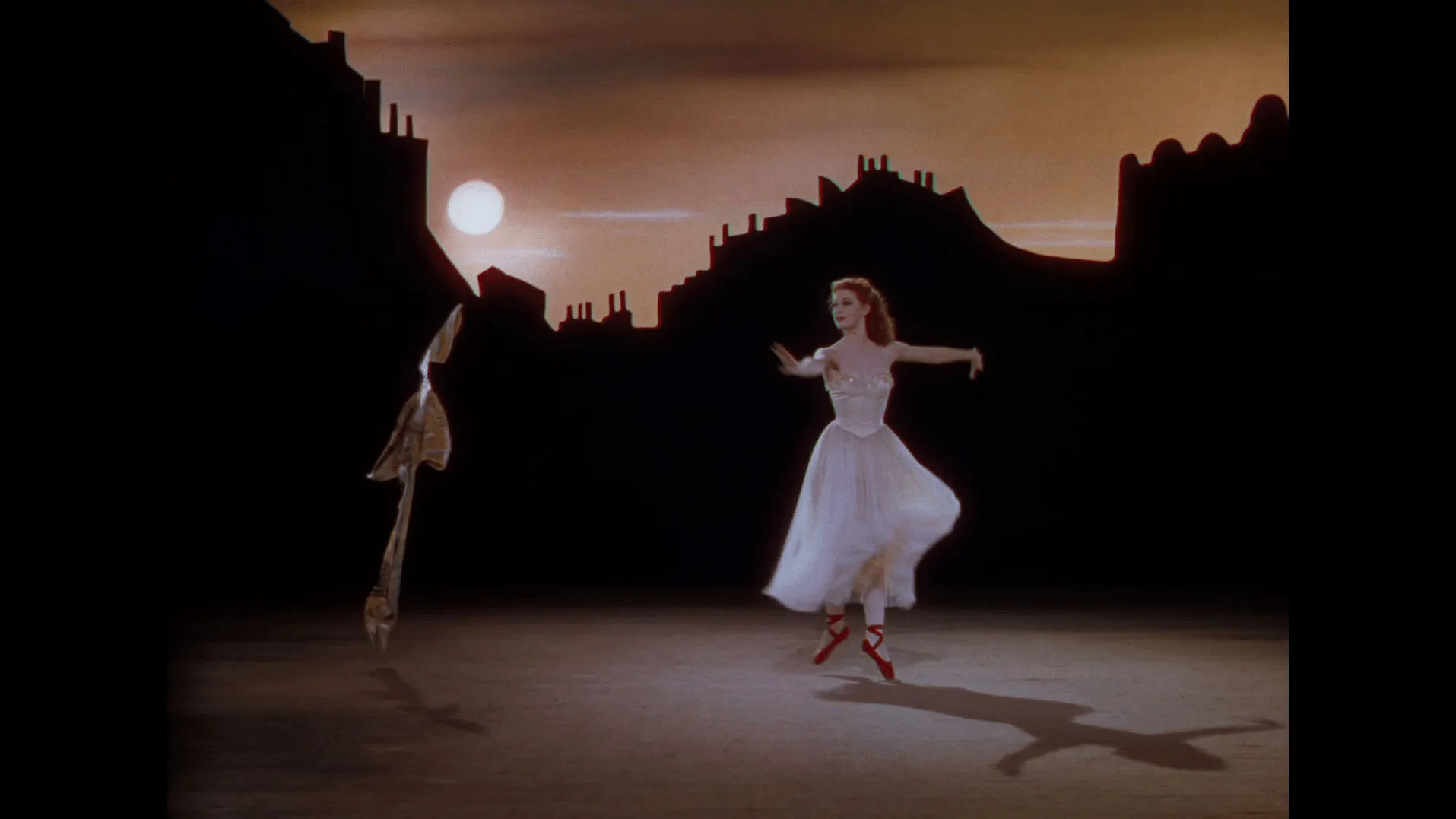 A white woman with red hair in a 40s style white ballet costume and red pointe shoes dancing with arms outstretched across a scene of a cityscape at dusk.