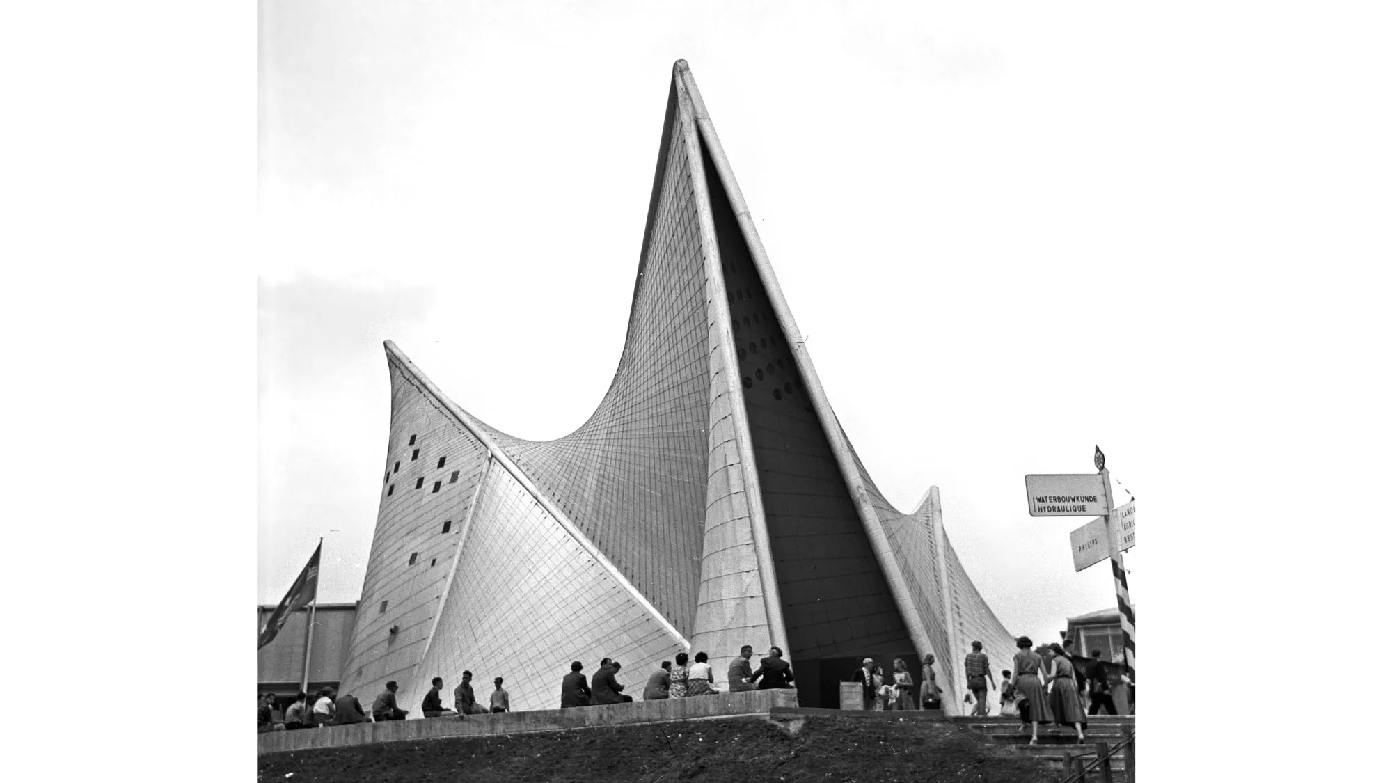 A man made cave type structure with a pointed roof and covered in glass. A small crowd looks on with backs to the camera. 