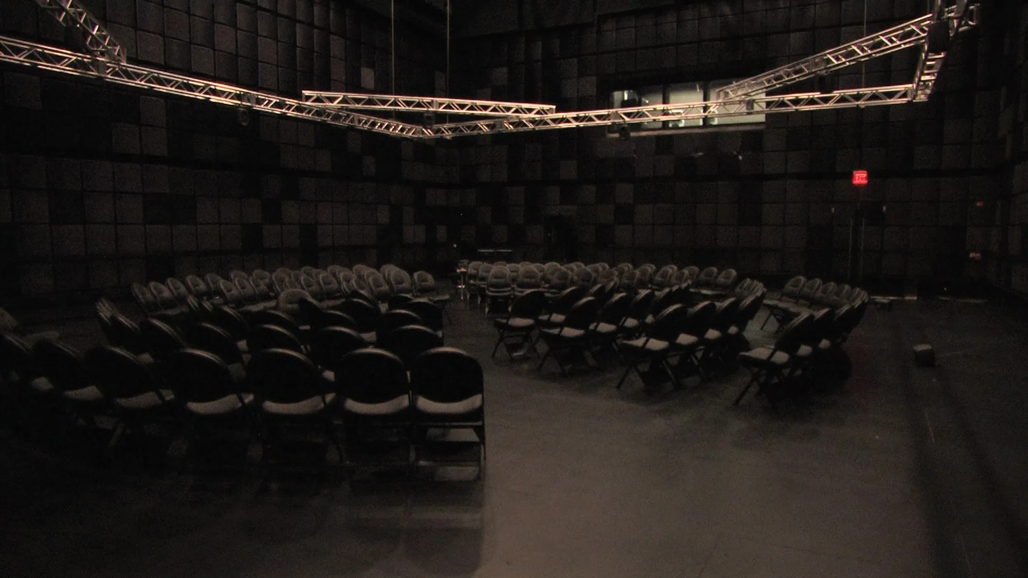 Many black folding chairs arranged in circle in a black box studio under a system of metal rigging suspended from the ceiling.