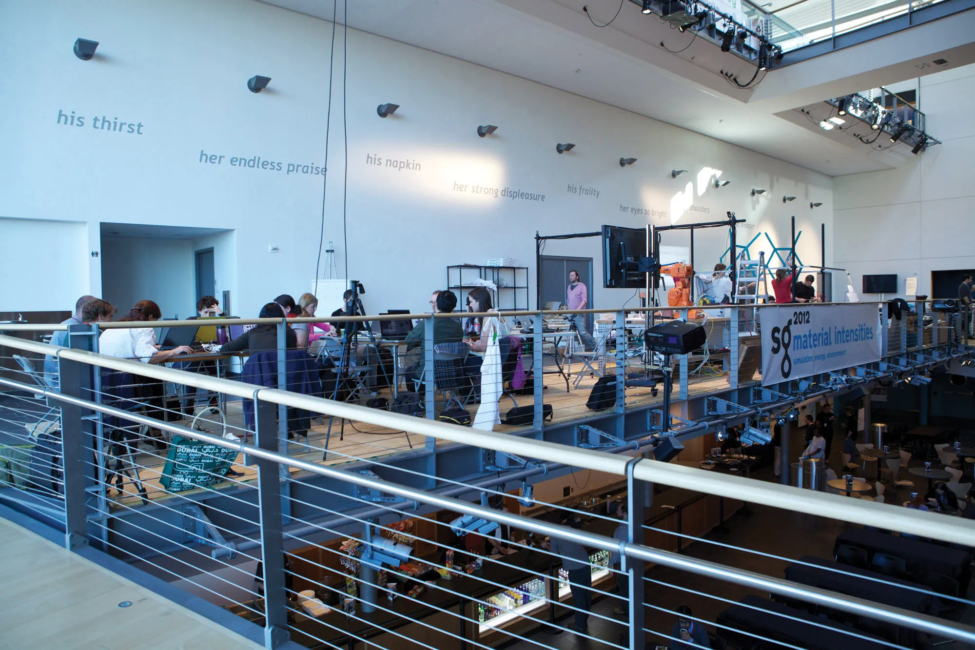 People seated at tables arranged in a mezzanine behind a gunmetal gray modern railing. On the white wall gray text reads, his thirst, her endless praise, his napkin. 